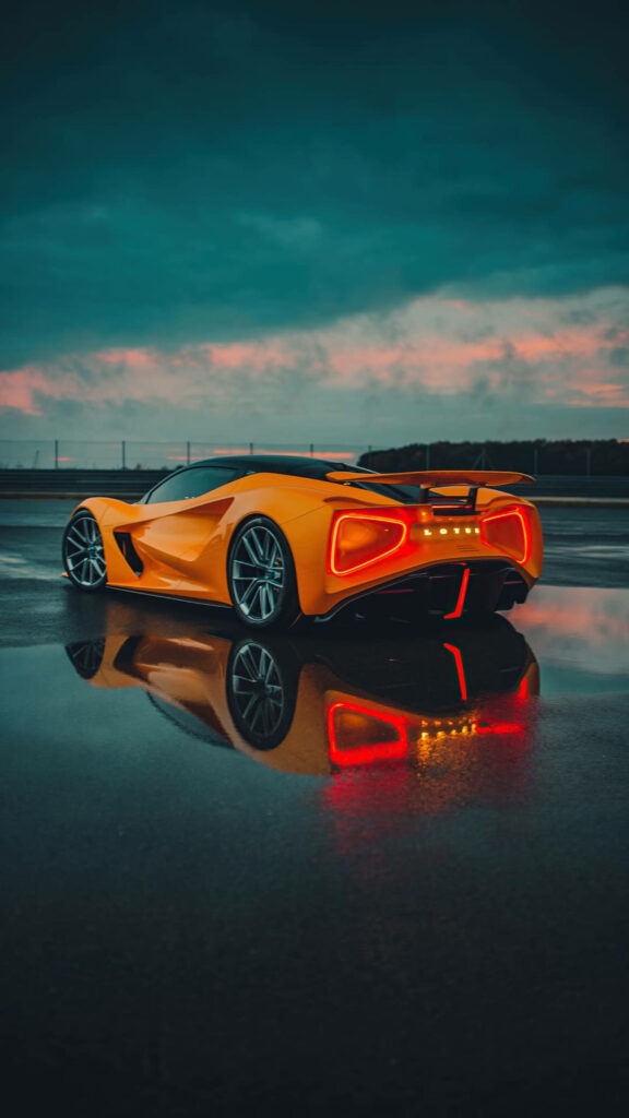 Orange sports car with glowing red tail lights reflected on wet surface under evening sky Wallpaper