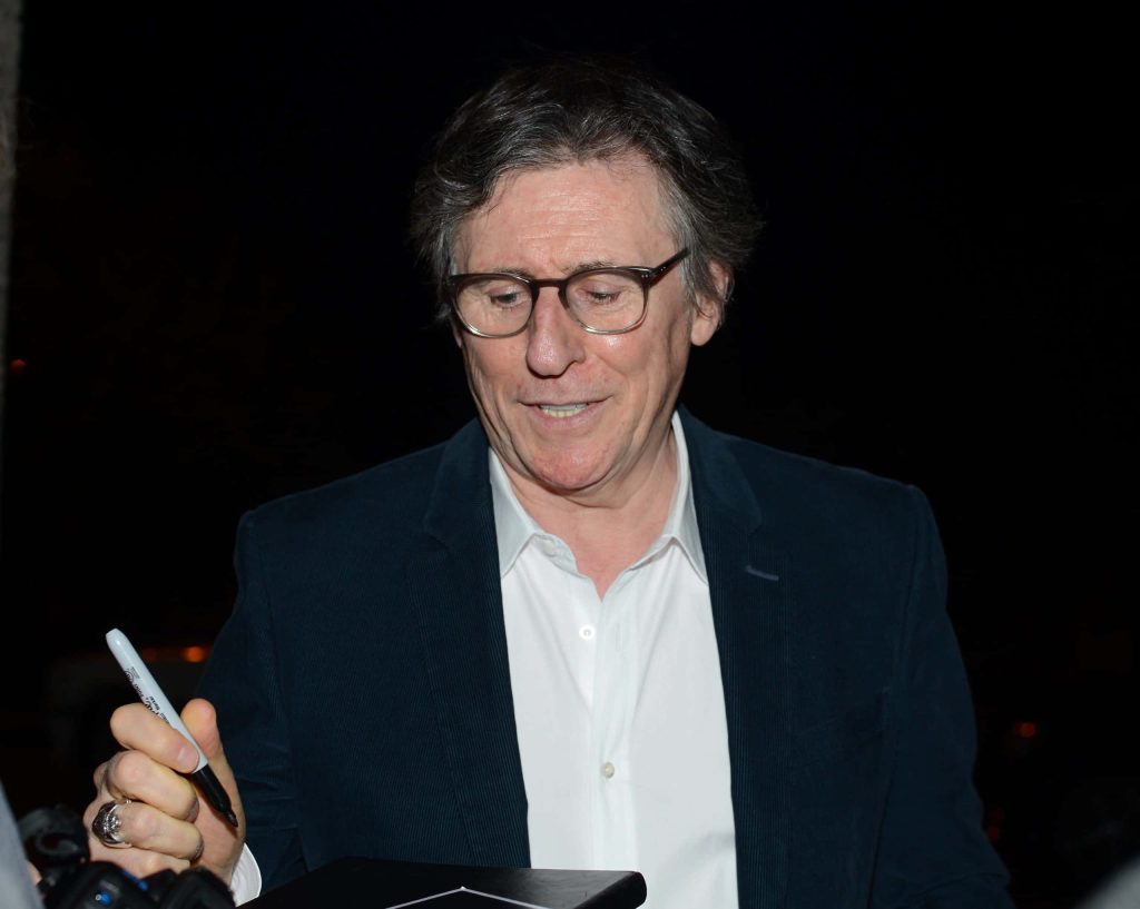 Gabriel Byrne signing autographs in sharp blazer white shirt dark moody wallpaper background