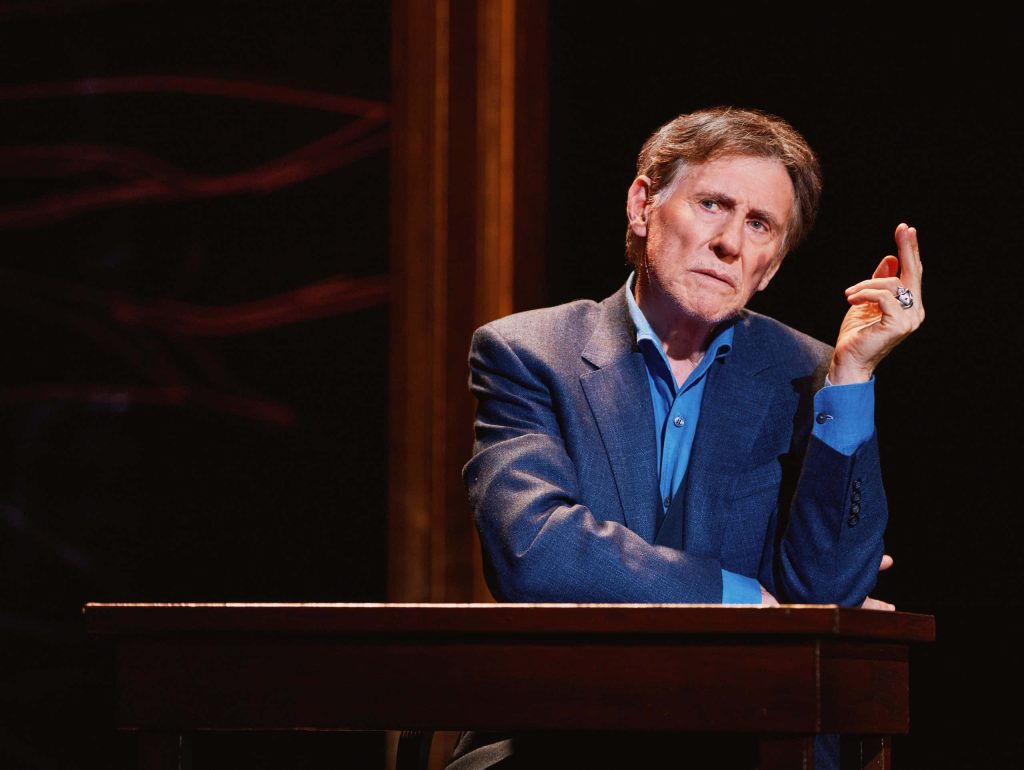 Gabriel Byrne portrait in blue blazer sitting at wooden table with dramatic dark background wallpaper