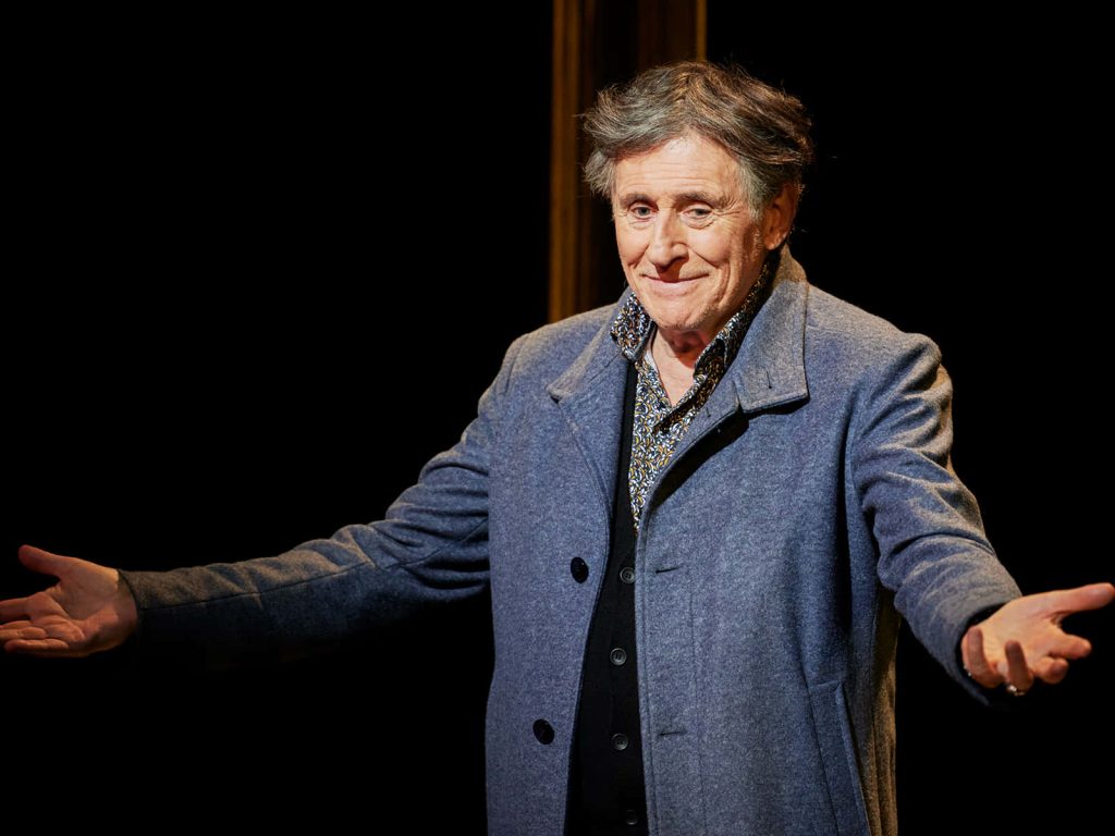Gabriel Byrne in warm theatrical light with open arms dark background iconic actor wallpaper