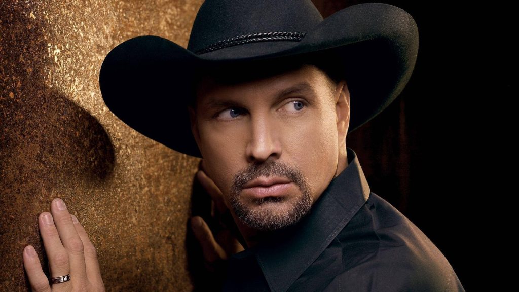 Garth Brooks in black cowboy hat and shirt leaning on rustic golden wall with dramatic lighting Wallpaper