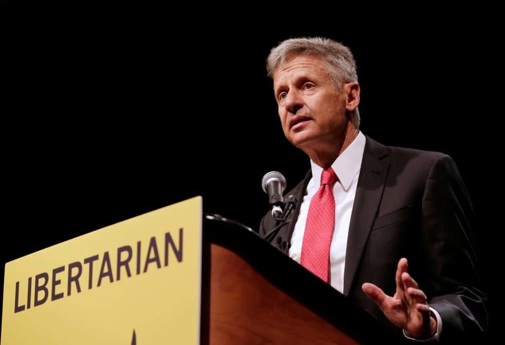 Gary Johnson speaking at podium in black suit red tie with yellow Libertarian sign background wallpaper