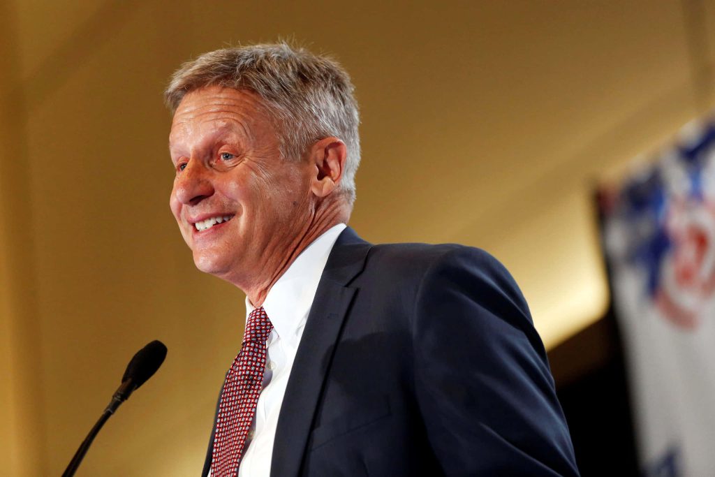 Gary Johnson smiling in dark suit red tie speaking at event with microphone yellow background wallpaper