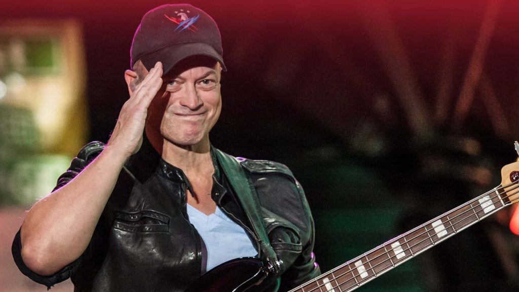 Gary Sinise performs live in leather jacket and cap saluting crowd with bass guitar on stage Wallpaper