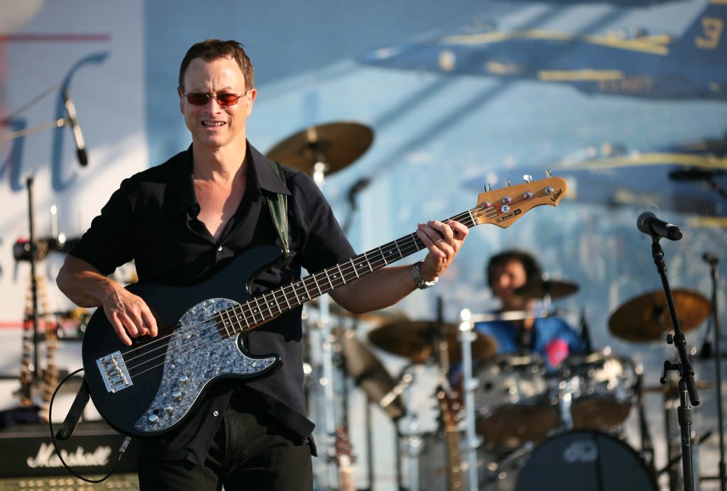 Gary Sinise playing black Line 6 bass guitar outdoors with drums and aircraft backdrop in sunlight Wallpaper
