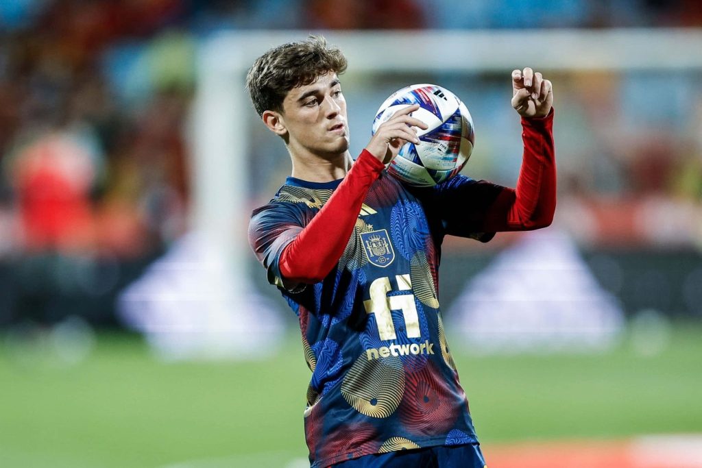 Spanish midfielder Gavi in Spain kit holding soccer ball on vibrant stadium background wallpaper