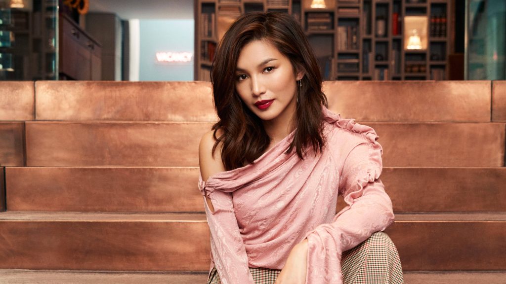 Gemma Chan seated on copper steps wearing pink ruffled top and plaid trousers in warm setting Wallpaper