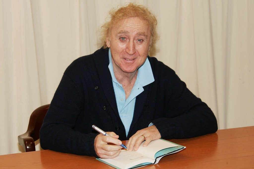 Gene Wilder smiling warmly signing book at wooden desk in cozy cardigan bright wallpaper background