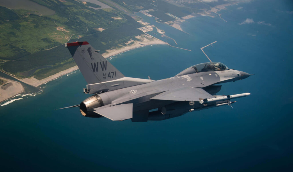 General Dynamics F-16 Fighting Falcon jet soaring over coastline with ocean and green landscape below Wallpaper