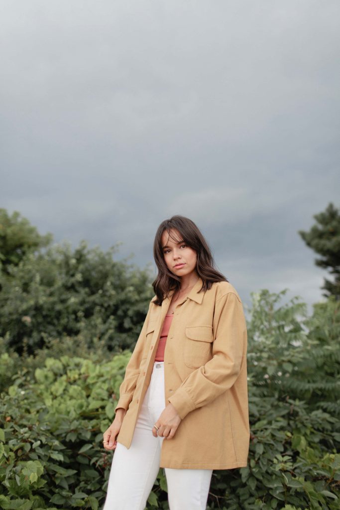 Genevieve Kang portrait in tan shacket white jeans lush greenery soft overcast sky wallpaper