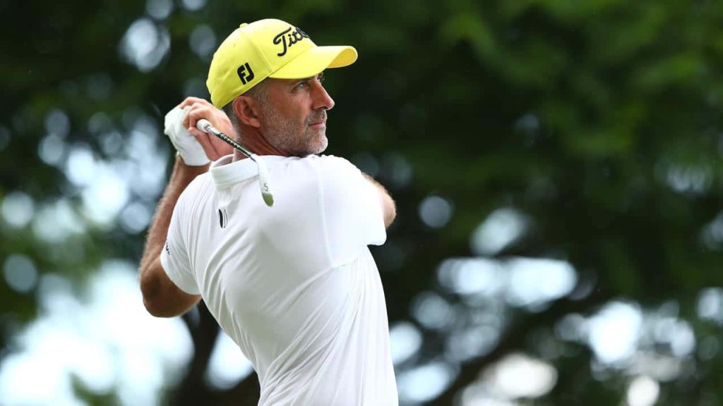 Professional golfer Geoff Ogilvy in yellow cap mid-swing with blurred green golf course background Wallpaper