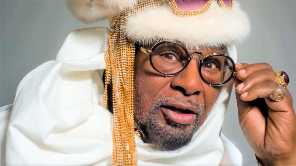 Close-up of George Clinton in white fur headpiece gold chains round glasses vibrant style wallpaper