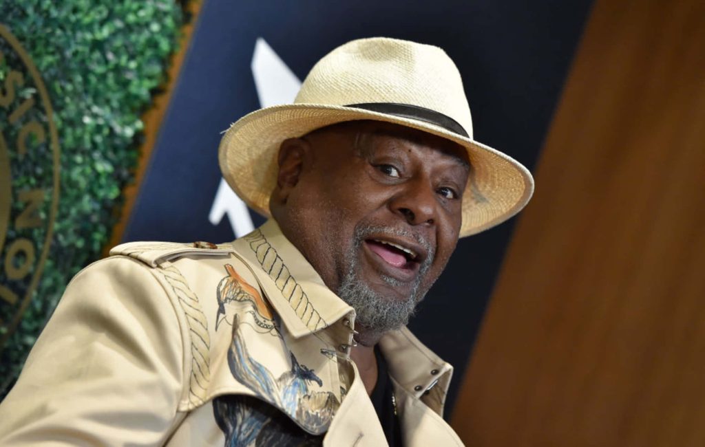 Portrait of funk legend George Clinton in straw fedora and patterned tan jacket smiling warmly Wallpaper