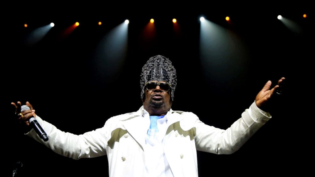 Funk legend George Clinton in white coat with avant-garde headpiece on vibrant concert stage Wallpaper