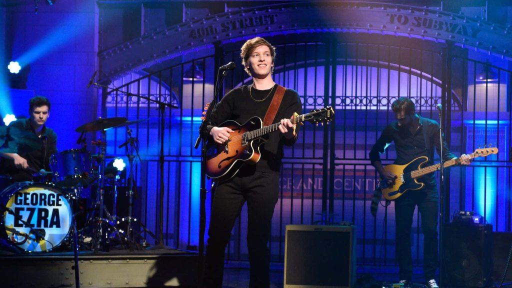 George Ezra live concert with blue lighting and subway backdrop playing guitar on stage Wallpaper