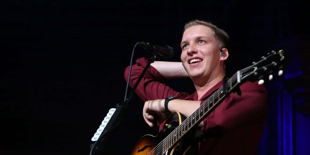 George Ezra live concert photo smiling playing guitar in maroon shirt dark moody stage background Wallpaper