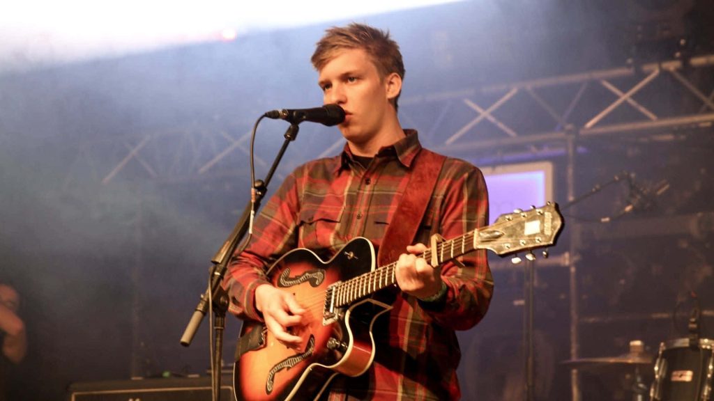 George Ezra performing live with archtop guitar and red plaid shirt on moody concert stage Wallpaper