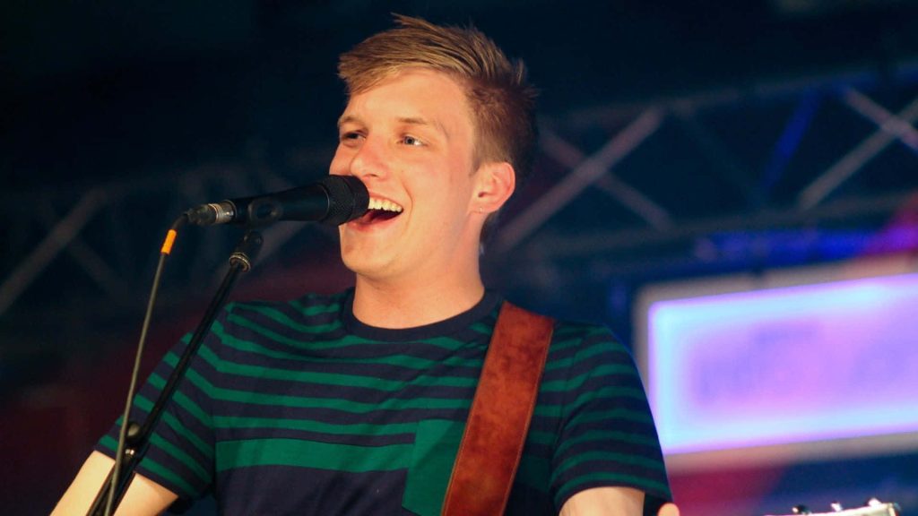 George Ezra singing live in navy green striped tee with vibrant stage lighting background wallpaper