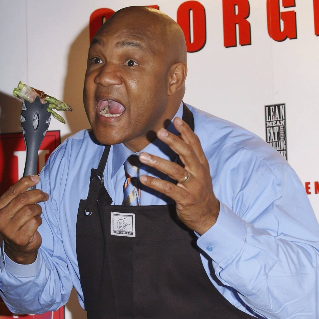 George Foreman in black apron holding tongs smiling with George Foreman Grill wallpaper background