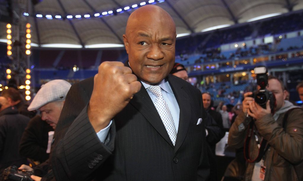 George Foreman in suit smiling and flexing fist in bright stadium celebrating boxing legacy wallpaper