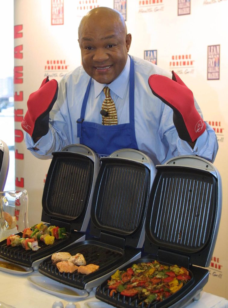 George Foreman in blue apron and red mittens grilling vegetables on multiple grills wallpaper