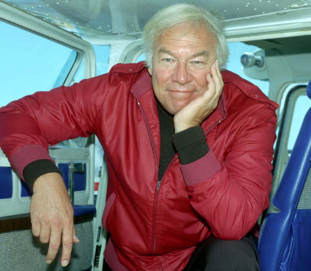 George Kennedy smiling in red jacket inside small aircraft candid wallpaper background