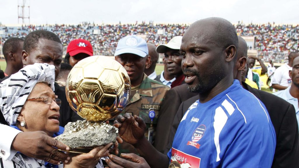 George Weah in blue jersey holding Ballon dOr trophy with Ellen Johnson Sirleaf stadium background Wallpaper