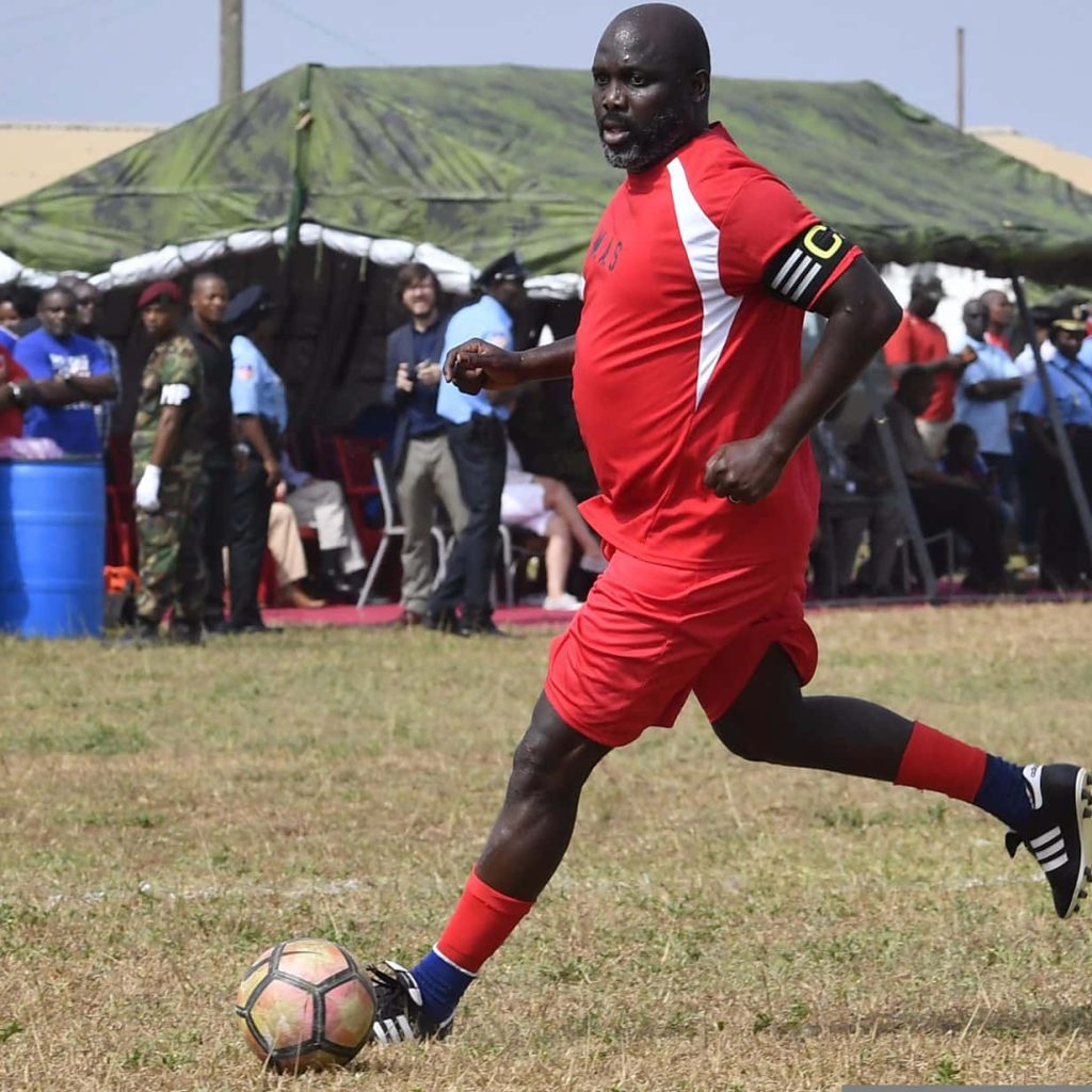 George Weah in red kit running with ball on outdoor field during friendly football match wallpaper