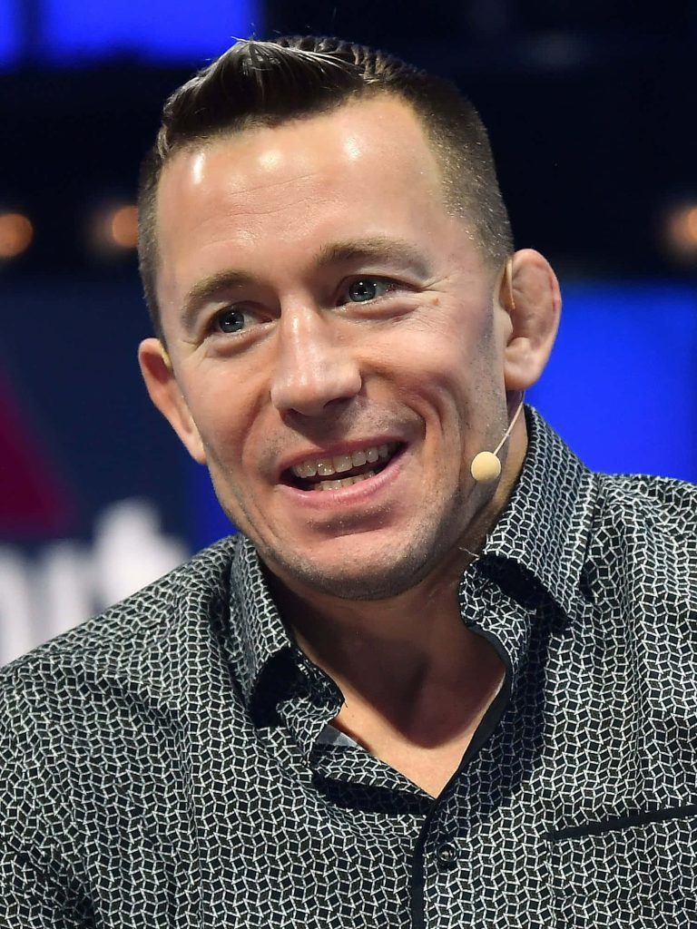Georges St-Pierre smiling in dark patterned shirt with lapel mic on soft blue background wallpaper
