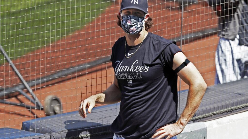 MLB pitcher Gerrit Cole in Yankees mask and tee behind dugout fence bright baseball field Wallpaper