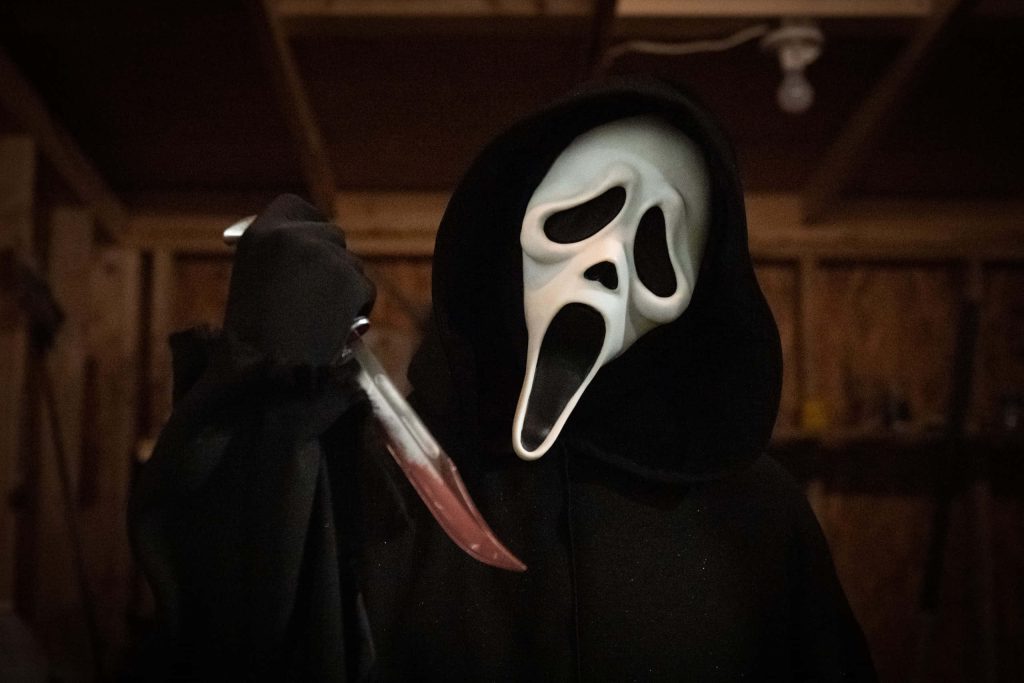 Ghostface from Scream in black robe holding blood-stained dagger in dim rustic horror setting Wallpaper