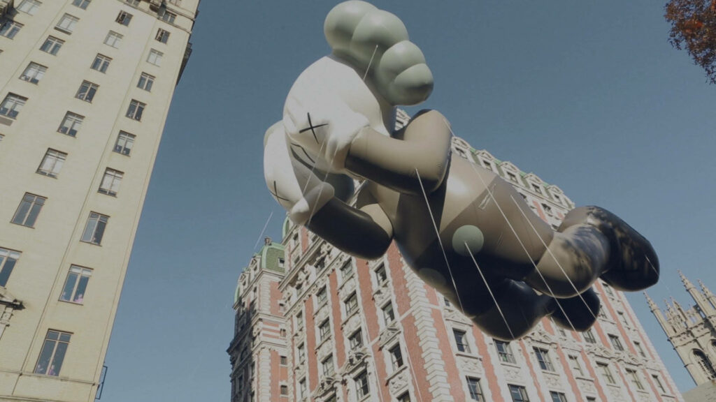 Giant KAWS Passing Through balloon floating between tall city buildings urban scene wallpaper