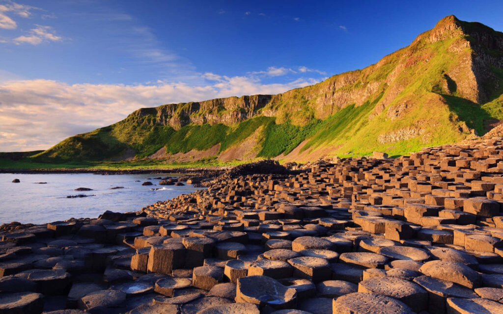 The Majestic Giants Causeway: A Spectacular Nature Preserve Snapshot with Towering Rock Formation and Mountain Silhouette, Bathed in Sunlight beneath a Hazy Azure Sky Wallpaper