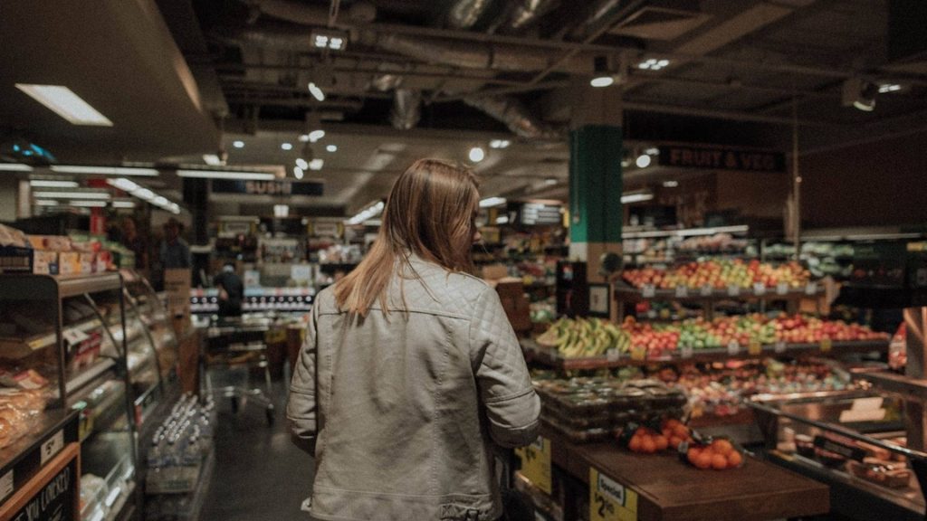Young woman in grey jacket shopping in grocery store aisle near fresh produce and sushi sign Wallpaper