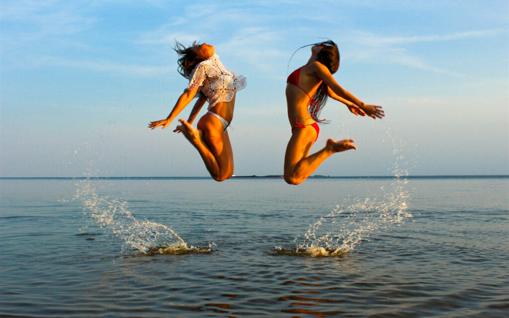 Two girls in colorful bikinis jump above shallow sea splashing water during vibrant sunset Wallpaper