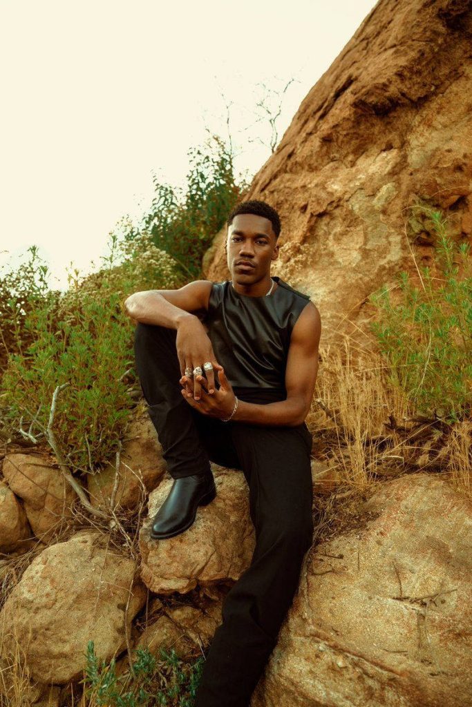 Giveon portrait sitting on sunlit rugged rocks in black leather top stylish outdoor wallpaper