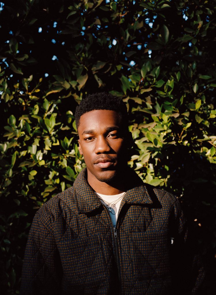 R&B singer Giveon in plaid jacket under dramatic natural light with lush green foliage background Wallpaper