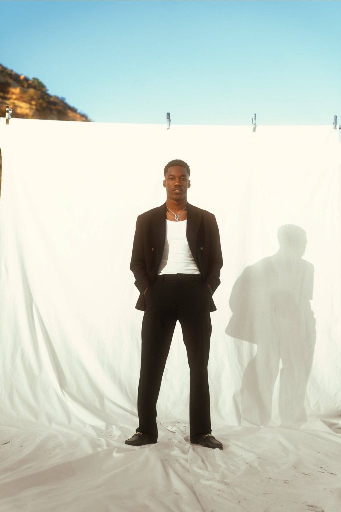 Giveon in black blazer and white tank top posing outdoors with dramatic sunlight shadow wallpaper