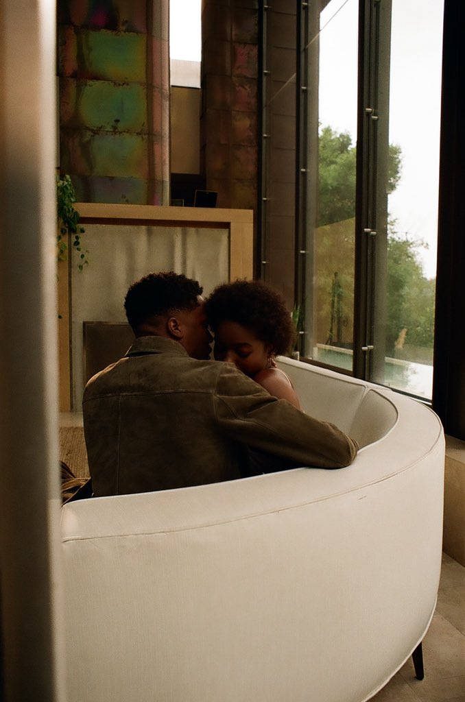 Giveon and partner embracing in white curved seating with romantic lighting and floor windows Wallpaper
