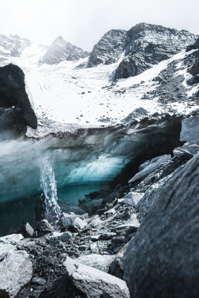 Alpine glacier cave with blue ice flowing water rugged rocks snowy peaks misty sky wallpaper background