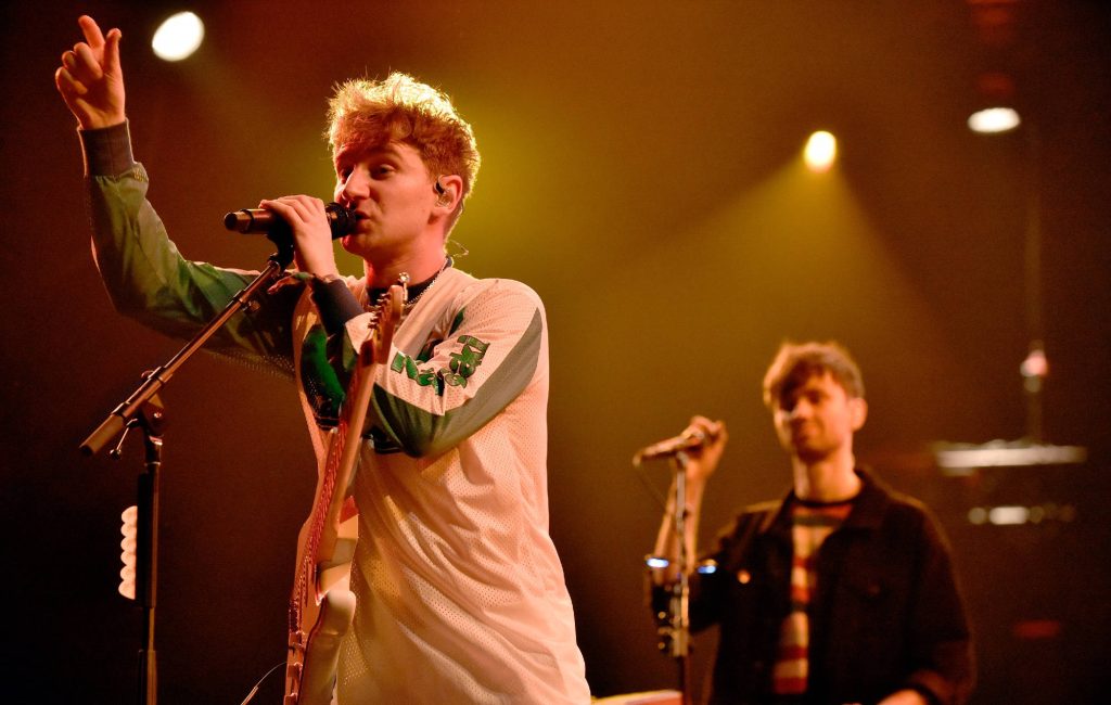 Glass Animals lead singer Dave Bayley performs electric guitar live under amber stage lights wallpaper