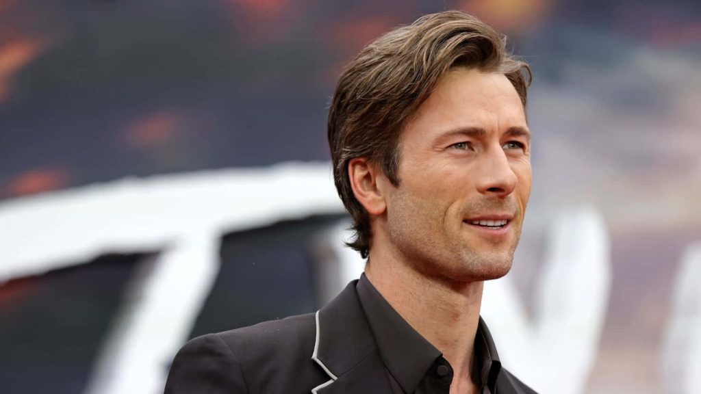 Glen Powell in dark suit smiling with styled hair bokeh event background professional wallpaper
