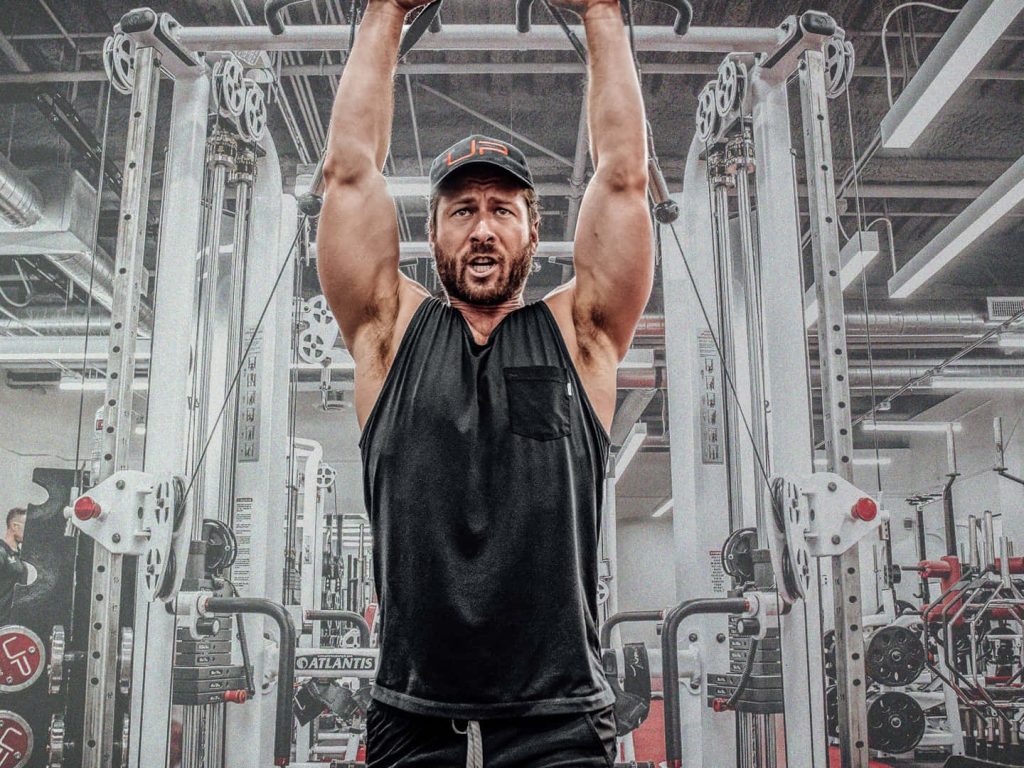 Glen Powell training with cable exercises in black tank top at Ultimate Performance gym intense workout Wallpaper