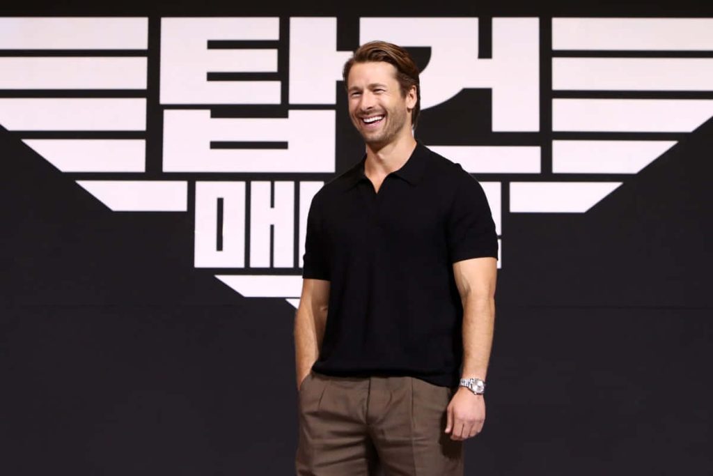 Glen Powell smiling in black polo and brown trousers at Top Gun Maverick promotional event background Wallpaper