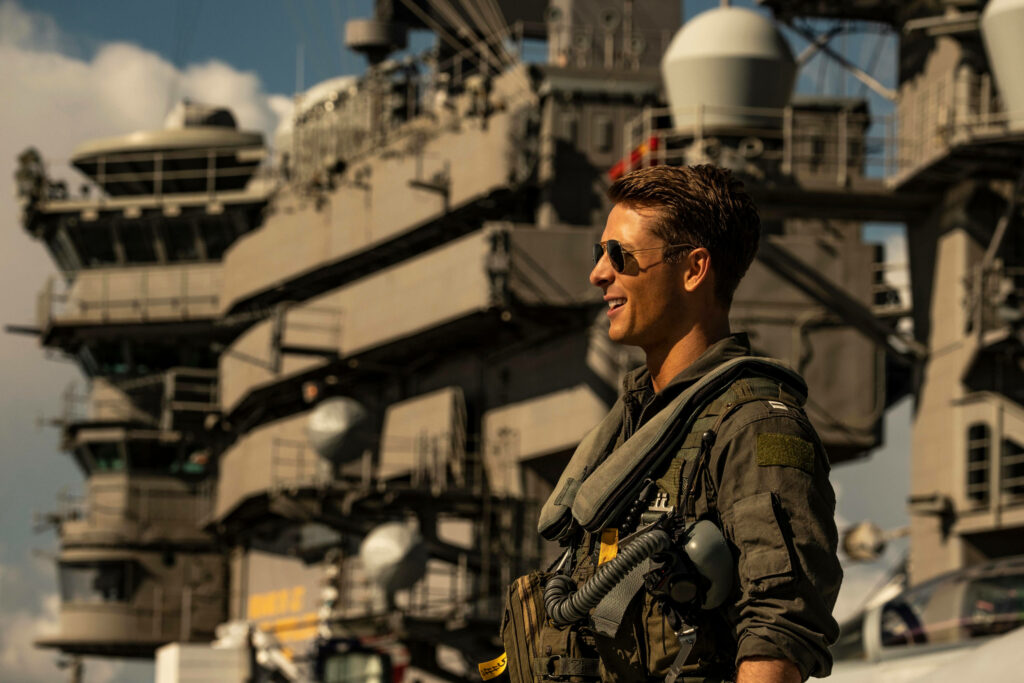 Glen Powell as Maverick flight suit stands confidently on aircraft carrier Top Gun Maverick wallpaper