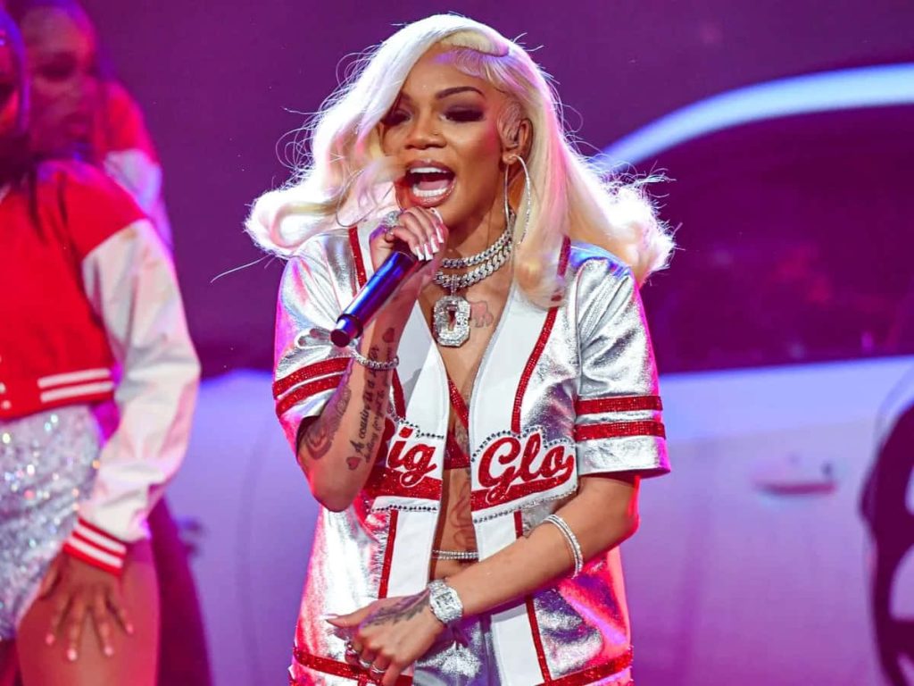 Rapper GloRilla in silver Big Glo jersey with platinum blonde hair on vibrant concert stage background Wallpaper