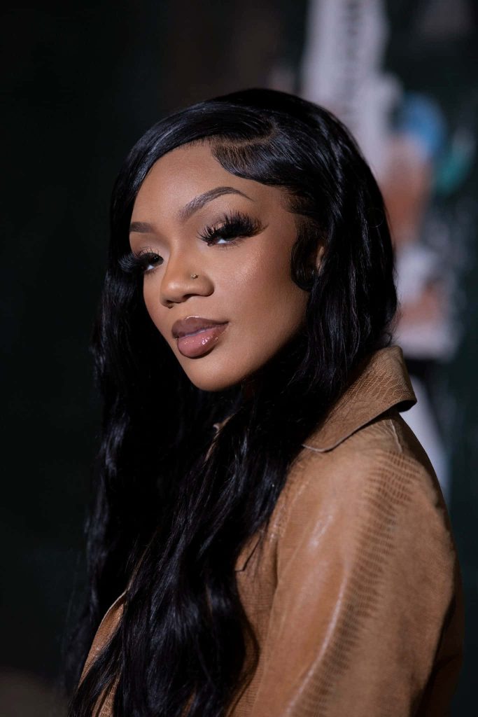 Portrait of rapper GloRilla with wavy black hair and glam makeup in tan jacket dark background Wallpaper