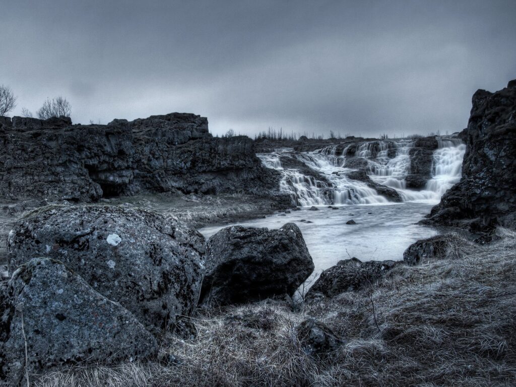 Gloomy landscape wallpaper with muted waterfall over rugged stones and faded grass under cloudy sky