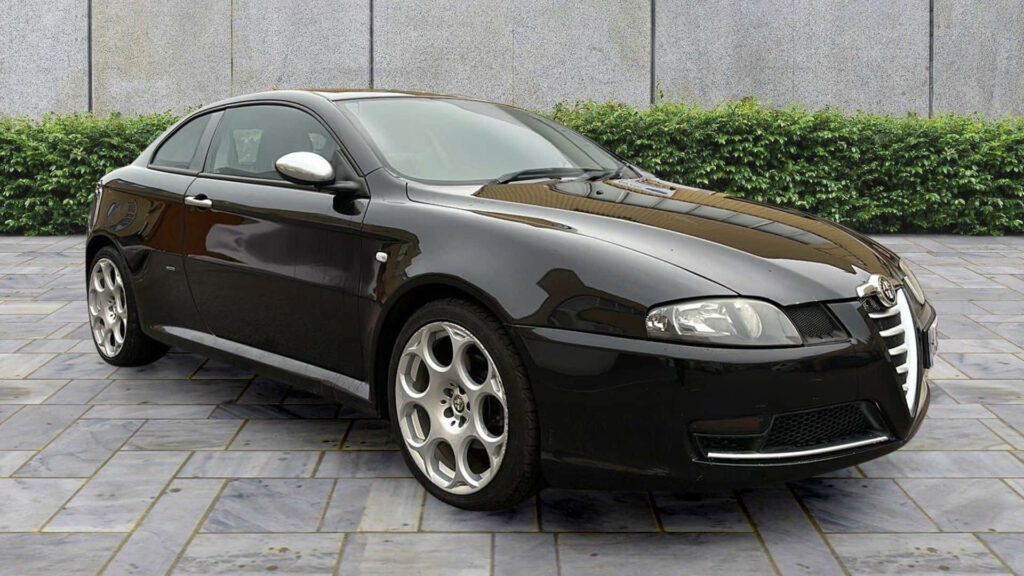 Glossy black Alfa Romeo GT parked on stone pavement with green foliage showcasing sporty design Wallpaper