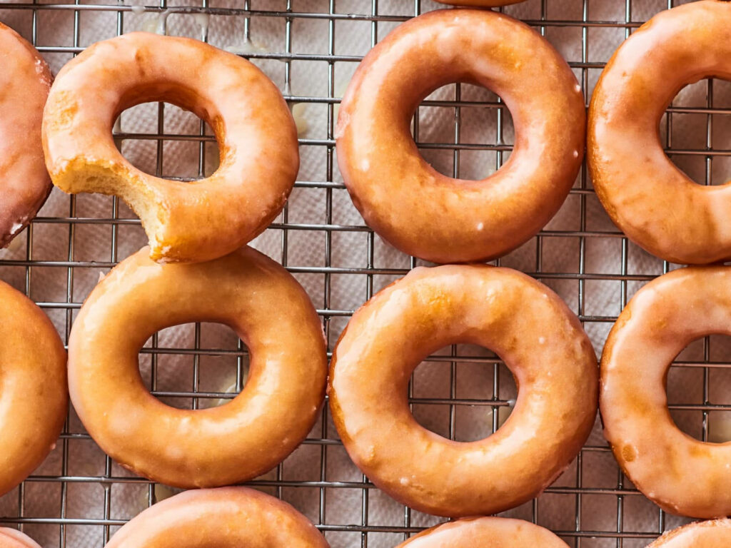 Rows of golden glazed donuts on cooling rack with one bitten donut vibrant food wallpaper